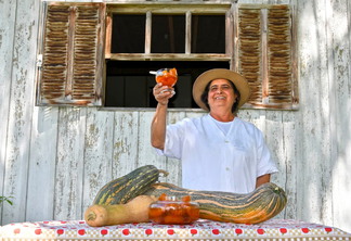 Foto de mulher em frente a uma janela. Ela sorri, segura uma bebida com a mão e está atrás de uma mesa com verduras.