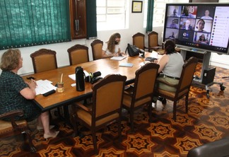 Foto de três mulheres em mesa de reunião. Em uma das pontas, uma TV apresenta uma videoconferência.