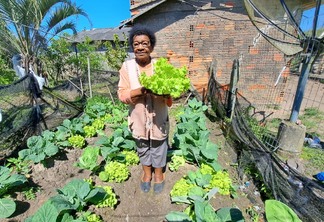 Foto de idosa negra segurando hortaliças em meio a uma horta.