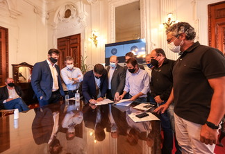 Foto de homens com máscara ao redor de mesa com papéis. O governador Eduardo Leite está assinando um deles.
