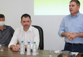 Foto de três homens em mesa de reunião. Um deles está falando em pé.