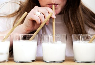 A foto mostra quatro copos com leite e uma mulher loira de casado branco, tomando leite através de um canudo.