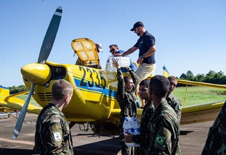 Foto de um grupo de militares alcançando água para dois homens estão colocando as garrafas em um avião agrícola amarelo.