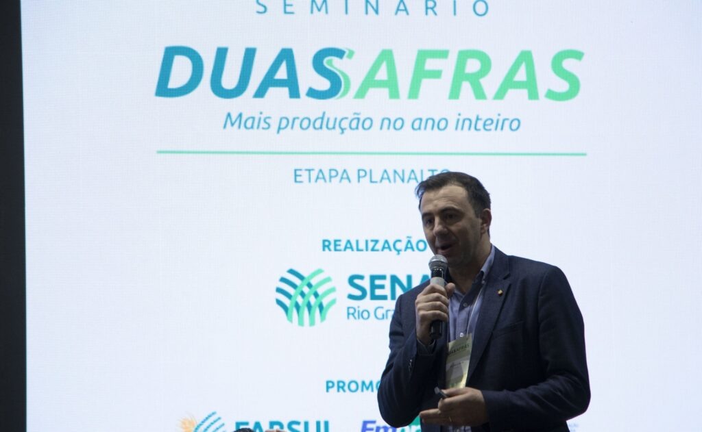 Foto de homem branco, com terno preto falando em microfone falando em frente a painel eletrônico do Seminário Duas Safras.,