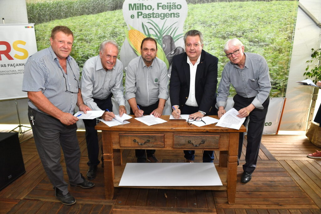Foto de cinco homens brancos assinando documentos em frente a banner do Programa Milho, Feijão e Pastagens.