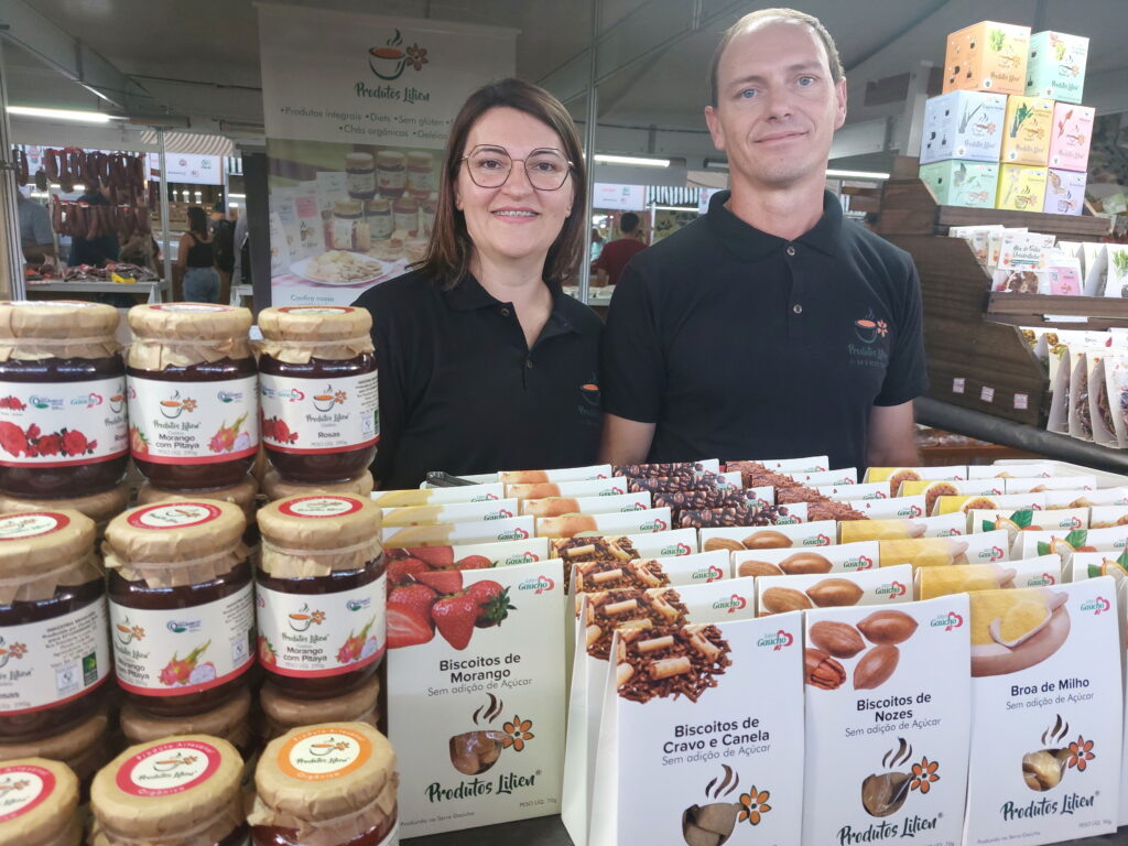 Foto de homem e mulher atrás de estande de geleias e biscoitos sorrindo.