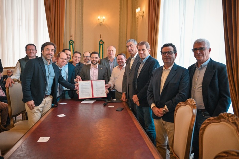 Foto do governador em exercício Gabriel Souza segurando um decreto aberto atrás de uma mesa. Ao redor dele estão diversos homens posando para foto.