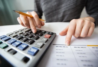 Foto focada em uma mão que está mexendo em uma calculadora enquanto segura um lápis e outra que está sobre papel com números.