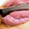 Foto de carne de frango sobre tábua de madeira. Uma faca está cortando a carne.