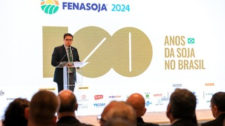 Foto de homem, com terno e gravata, falando em parlatório em um palco. Atrás dele está um telão anunciando a Fenasoja 2024 - 100 Anos da Soja no Brasil.