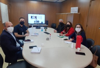 Foto de pessoas com máscaras ao redor de mesa de reuniões.