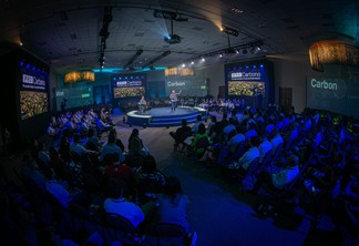 Foto de evento com pessoas sentadas ao redor de palco, onde estão dois homens. Em painéis ao redor está escrito "PRO Carbono". Foto de evento com pessoas sentadas ao redor de palco, onde estão dois homens. Em painéis ao redor está escrito "PRO Carbono".