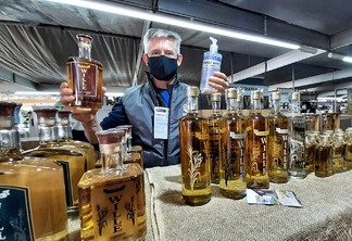 Foto de homem atrás de bancada de produtos. Ele segura uma garrafa de bebida em uma mão e um álcool em gel em outra.
