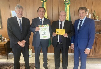 Foto de quatro homens posando para foto. No meio, Jair Bolsonaro e Nei César Manica seguram panfletos da Expodireto. Nenhum deles usa máscara.