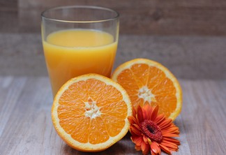 A foto mostra uma laranja cortada em duas partes, apoiadas em um copo de vidro com suco da fruta dentro.