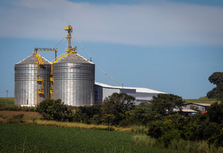 Imagem mostra silos de armazenamento de grãos ao fundo e em primeiro plano, uma lavoura e arvores nativas