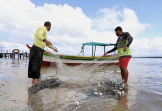 Imagem mostra dois homens mexendo em rede de pesca na praia