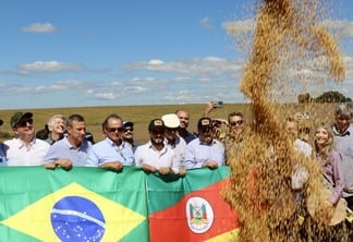 A foto mostra algumas pessoas segurando a bandeira do Rio Grande do Sul e do Brasil, e com soja caindo