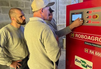 Foto de três homens em frente a uma máquina. Um deles está com a mão numa tela. A máquina tem a inscrição "Roboagro".