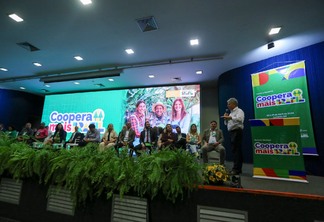 Foto de palco com várias pessoas sentadas enquanto o Ministro Paulo Teixeira, em pé, discursa. O telão no fundo e banners anunciam o "Programa Coopera Mais Brasil".
