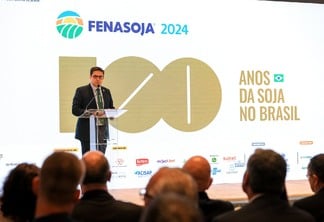 Foto de homem, com terno e gravata, falando em parlatório em um palco. Atrás dele está um telão anunciando a Fenasoja 2024 - 100 Anos da Soja no Brasil.