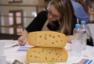 Foto de queijos empilhadas sobre mesa. Ao fundo uma mulher está sentada a mesa e escrevendo em um papel.