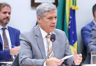 Foto de ministro Paulo Teixeira falando em microfone. Ele é branco, tem cabelo grisalho e usa terno cinza com gravata cinza e laranja.