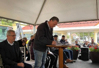 Foto de ministro Carlos Fávaro em pé enquanto assina papéis que estão sobre uma mesa. Ele está embaixo de uma lona de plástico e várias pessoas estão sentadas atrás dele. Foto de ministro Carlos Fávaro em pé enquanto assina papéis que estão sobre uma mesa. Ele está embaixo de uma lona de plástico e várias pessoas estão sentadas atrás dele.