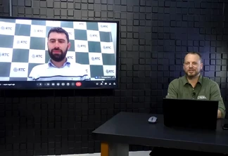 Foto com um homem branco sentado atrás de mesa. Ele usa uma camisa verde e tem um notebook, mouse e celular na sua frente. Ao lado dele, a TV mostra um homem branco com cabelo e barba castanhos e camisa branca. Foto com um homem branco sentado atrás de mesa. Ele usa uma camisa verde e tem um notebook, mouse e celular na sua frente. Ao lado dele, a TV mostra um homem branco com cabelo e barba castanhos e camisa branca.