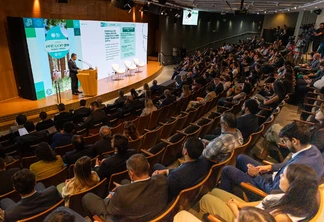 Foto de auditório com pessoas sentadas assistindo enquanto homem de terno fala em parlatório no palco em frente a um telão. Foto de auditório com pessoas sentadas assistindo enquanto homem de terno fala em parlatório no palco em frente a um telão.