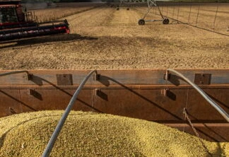 Imagem mostra um caminhão com grãos de soja, uma colheitadeira e um pivô de irrigação em uma lavoura de soja