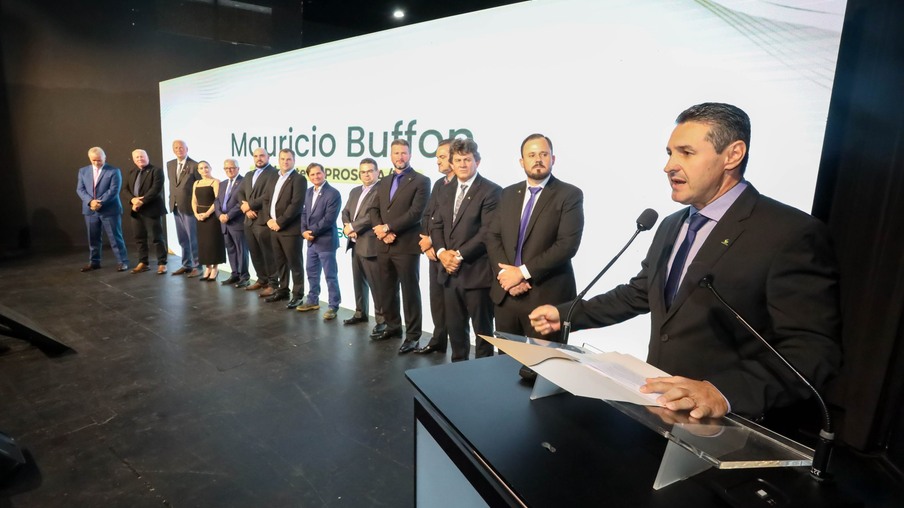 Foto de Maurício Buffon discursando em palco. Atrás dele está uma fila com vários homens e uma mulher. Foto de Maurício Buffon discursando em palco. Atrás dele está uma fila com vários homens e uma mulher.