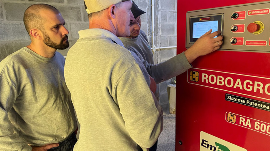 Foto de três homens em frente a uma máquina. Um deles está com a mão numa tela. A máquina tem a inscrição "Roboagro". Foto de três homens em frente a uma máquina. Um deles está com a mão numa tela. A máquina tem a inscrição "Roboagro".