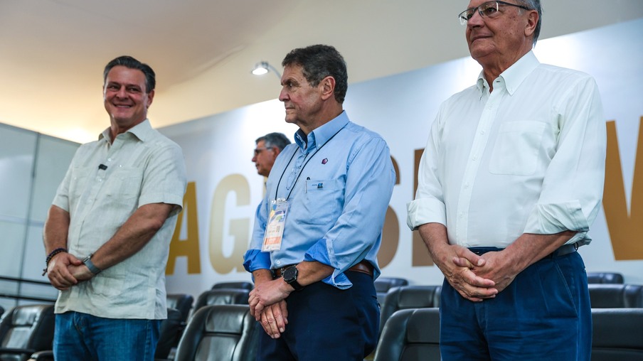 Foto de três homens em pé em um palco com as mãos cruzadas na frente do corpo. Foto de três homens em pé em um palco com as mãos cruzadas na frente do corpo.
