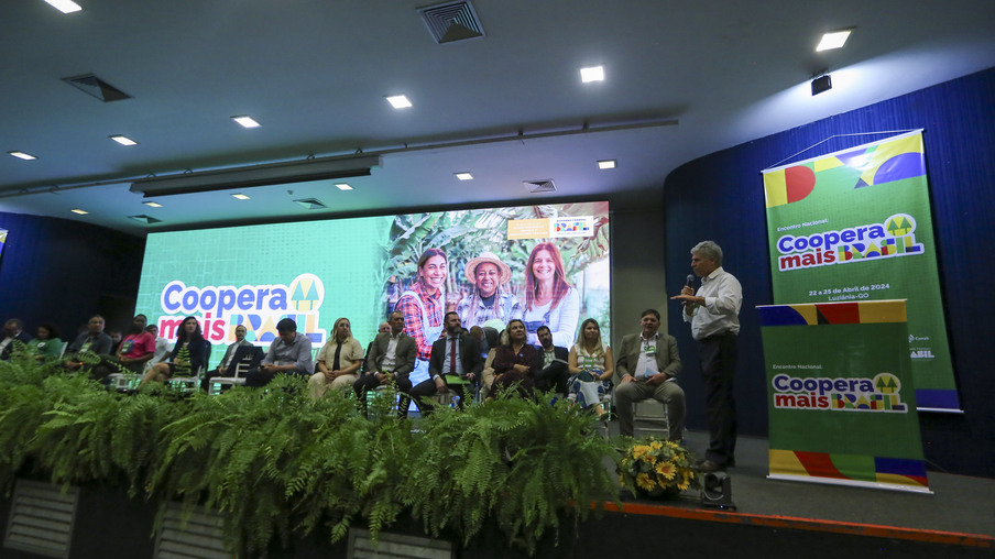 Foto de palco com várias pessoas sentadas enquanto o Ministro Paulo Teixeira, em pé, discursa. O telão no fundo e banners anunciam o "Programa Coopera Mais Brasil".
