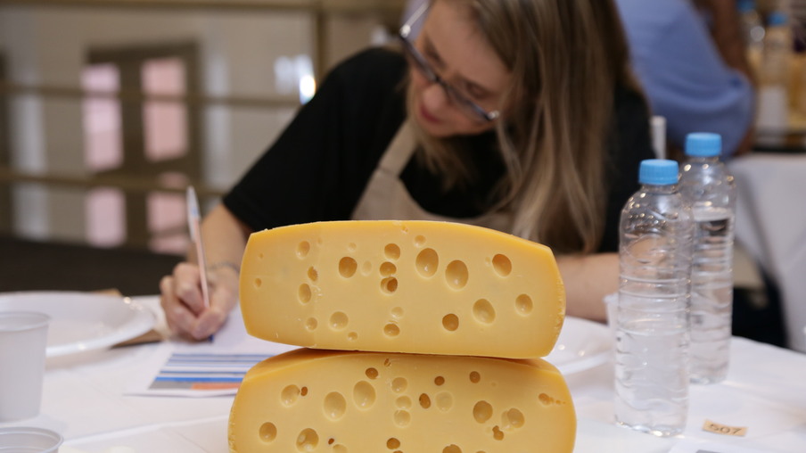 Foto de queijos empilhadas sobre mesa. Ao fundo uma mulher está sentada a mesa e escrevendo em um papel. Foto de queijos empilhadas sobre mesa. Ao fundo uma mulher está sentada a mesa e escrevendo em um papel.