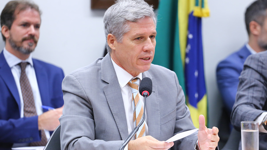Foto de ministro Paulo Teixeira falando em microfone. Ele é branco, tem cabelo grisalho e usa terno cinza com gravata cinza e laranja. Foto de ministro Paulo Teixeira falando em microfone. Ele é branco, tem cabelo grisalho e usa terno cinza com gravata cinza e laranja.