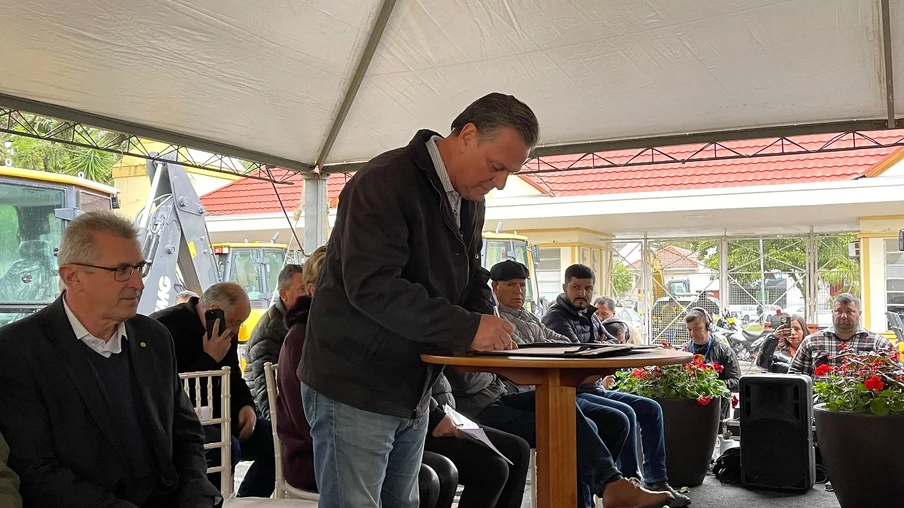 Foto de ministro Carlos Fávaro em pé enquanto assina papéis que estão sobre uma mesa. Ele está embaixo de uma lona de plástico e várias pessoas estão sentadas atrás dele. Foto de ministro Carlos Fávaro em pé enquanto assina papéis que estão sobre uma mesa. Ele está embaixo de uma lona de plástico e várias pessoas estão sentadas atrás dele.