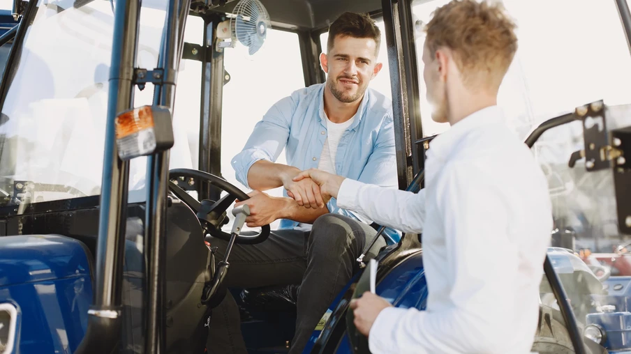 A foto mostra dois homens, um deles está dentro da cabine de um trator e o outro está do lado de fora. Eles estão dando um cumprimento de mãos. A foto mostra dois homens, um deles está dentro da cabine de um trator e o outro está do lado de fora. Eles estão dando um cumprimento de mãos.