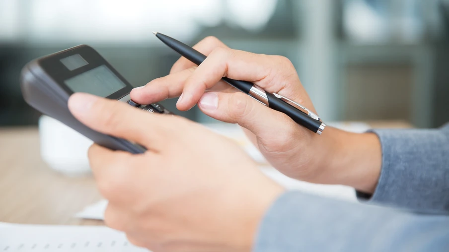 A foto mostra mãos segurando e digitando em uma calculadora enquanto seguram uma caneta. A foto mostra mãos segurando e digitando em uma calculadora enquanto seguram uma caneta.
