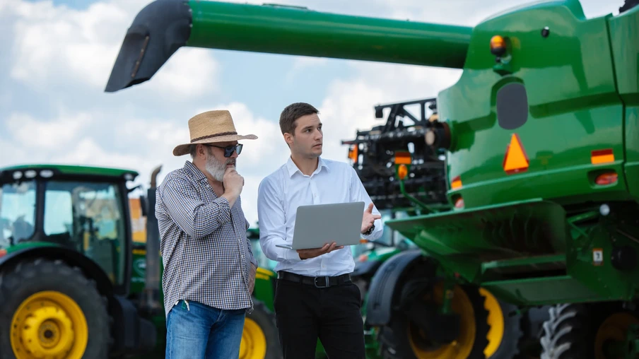 Foto de dois homens ao lado de máquina agrícola verde olhando para o notebook que um deles segura. Foto de dois homens ao lado de máquina agrícola verde olhando para o notebook que um deles segura.