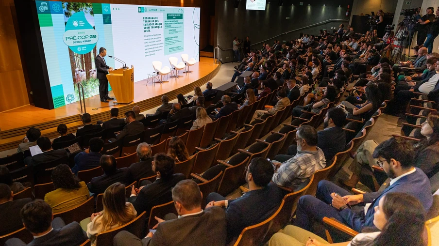 Foto de auditório com pessoas sentadas assistindo enquanto homem de terno fala em parlatório no palco em frente a um telão. Foto de auditório com pessoas sentadas assistindo enquanto homem de terno fala em parlatório no palco em frente a um telão.
