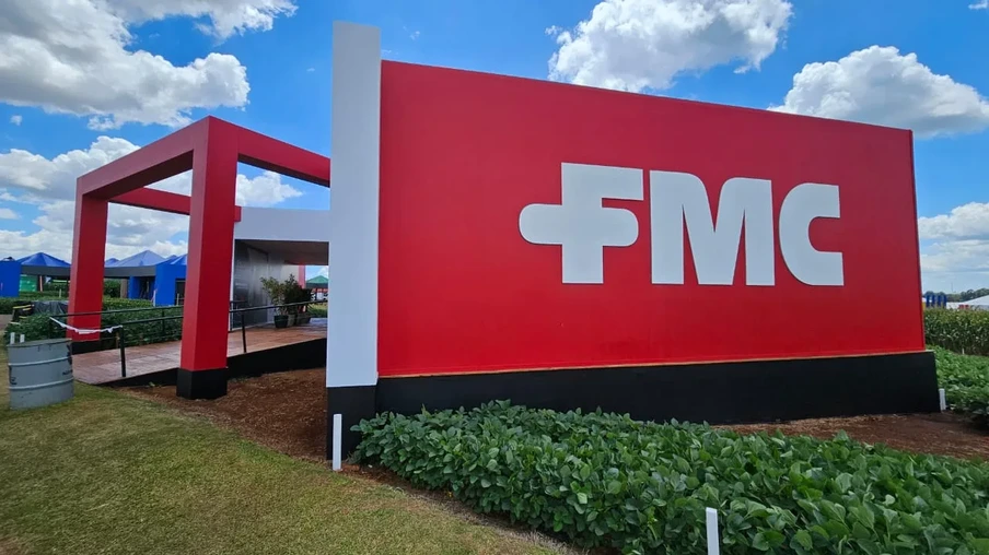 Foto de estande da FMC. Ele tem um painel grande vermelho com FMC escrito em branco. Na frente há plantas de soja e ao fundo está a entrada do estande. Foto de estande da FMC. Ele tem um painel grande vermelho com FMC escrito em branco. Na frente há plantas de soja e ao fundo está a entrada do estande.