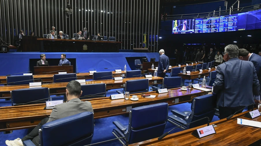 Foto do Plenário do Senado Federal. Em primeiro plano estão as mesas com pessoas sentadas e em pé atrás delas. No fundo está a mesa diretora do Senado e painéis. Foto do Plenário do Senado Federal. Em primeiro plano estão as mesas com pessoas sentadas e em pé atrás delas. No fundo está a mesa diretora do Senado e painéis.