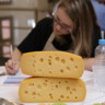 Foto de queijos empilhadas sobre mesa. Ao fundo uma mulher está sentada a mesa e escrevendo em um papel.
